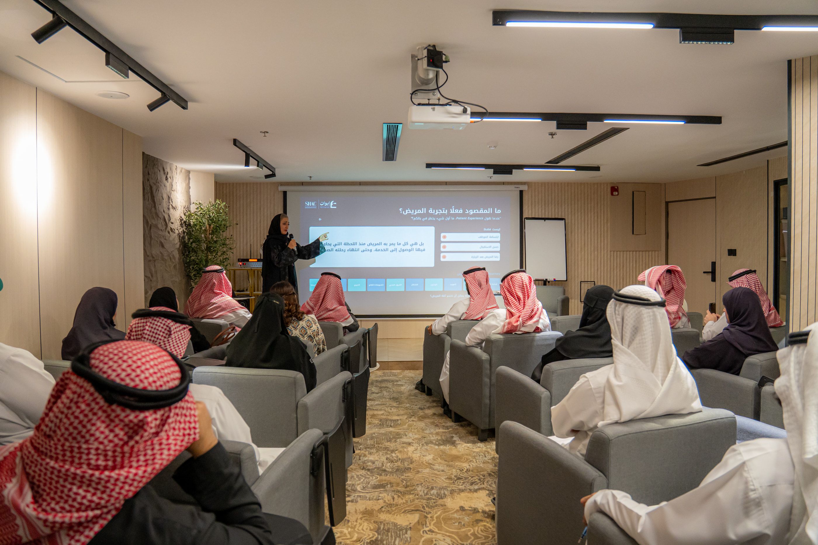 قاعة الجلسة من الخلف: الحضور أمام المتحدثة وشريحة «ما المقصود فعلًا بتجربة المريض؟»
