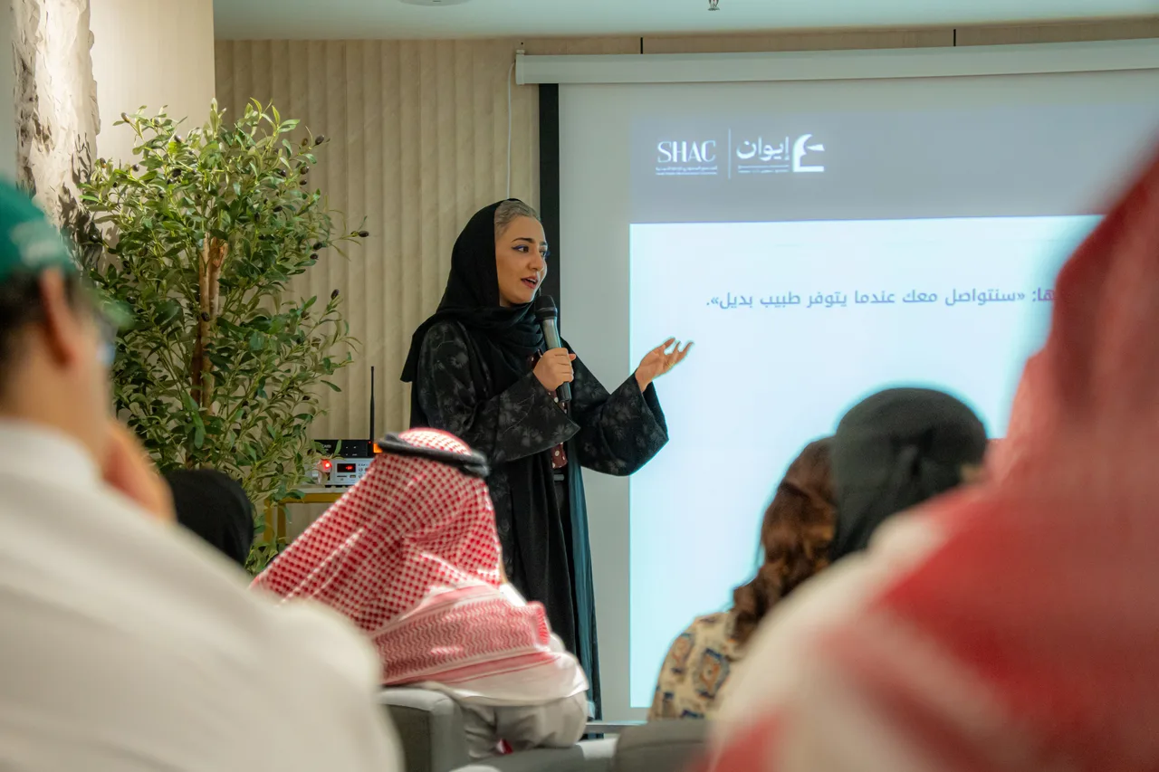 المتحدثة في جلسة إيوان أمام شريحة تحمل شعارَي SHAC وإيوان وعبارة «سنتواصل معك عندما يتوفر طبيب بديل»، والحضور من الخلف