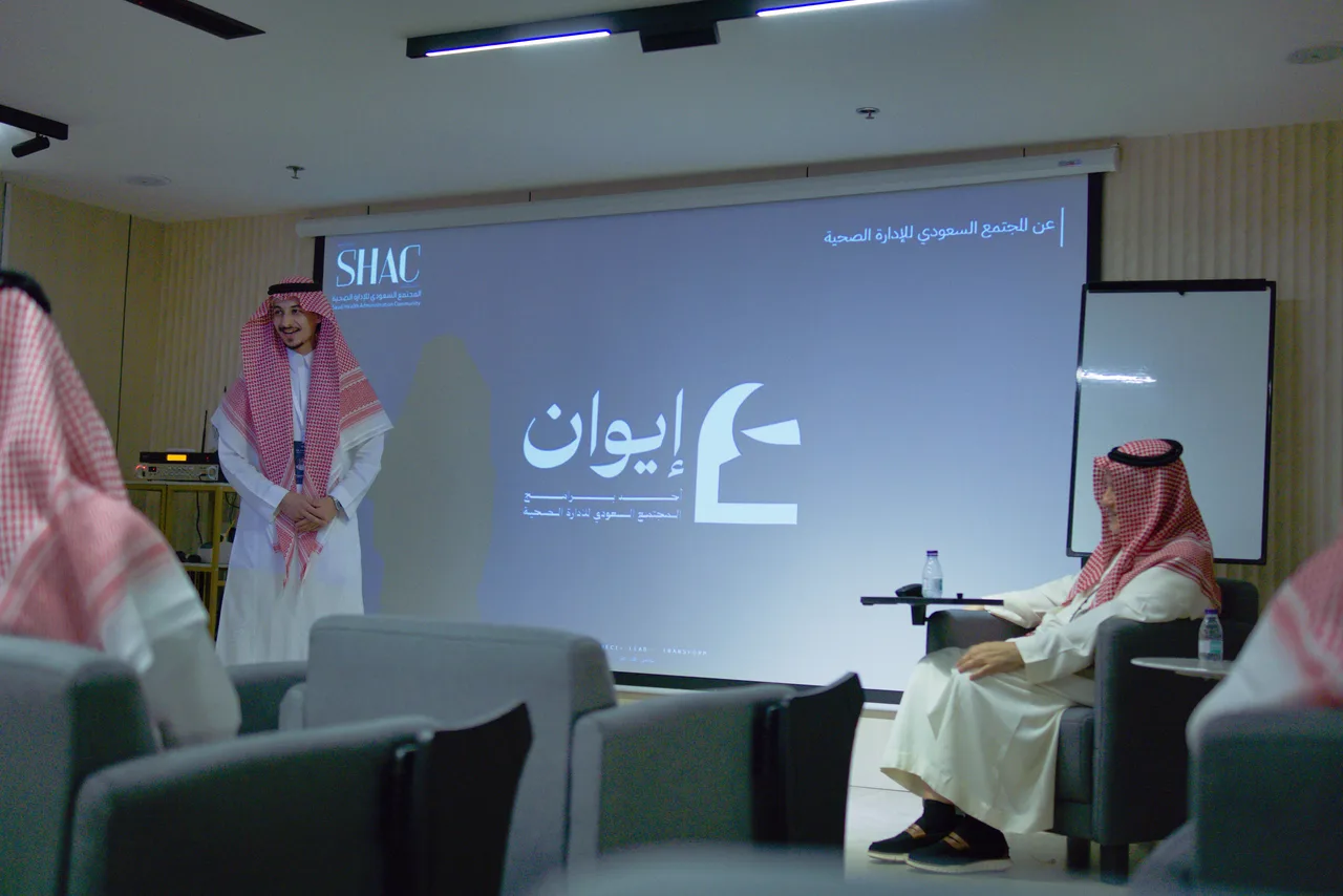 لقاء إيوان من آخر القاعة: شريحة العنوان تحمل شعار إيوان، ومقدّم اللقاء واقف، والضيف جالس، والحضور من الخلف