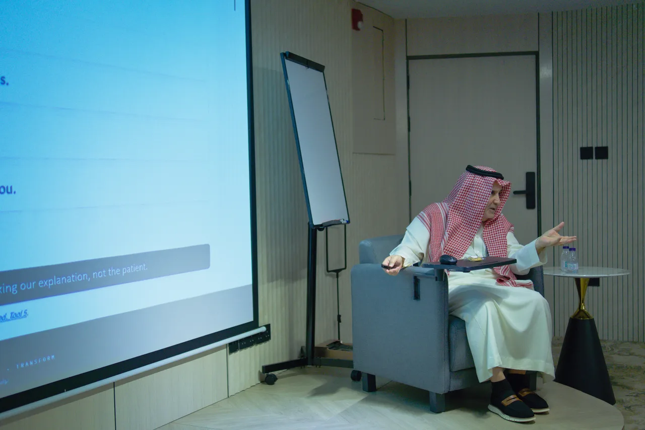 مقدّم اللقاء جالسًا بجوار الشاشة يخاطب الحضور
