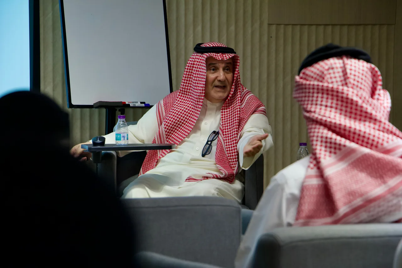 مقدّم لقاء إيوان يتحدث ويده مفتوحة، بين اثنين من الحضور